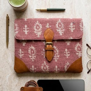 Pode incluir: Uma capa para tablet rosa empoeirado com um padrão creme e detalhes em couro. A capa tem um fecho de fivela de couro e cantos de couro. Uma caneta dourada, uma xícara de matcha e óculos também são visíveis.