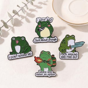 Könnte beinhalten: Vier grüne Frosch-Emaille-Anstecknadeln mit schwarzen Umrissen und weißen Akzenten. Jede Anstecknadel zeigt ein anderes Froschdesign mit Text: "I DON'T WANNA MO", "I have had Just about enough", "FUCK AROUND AND FIND OUT", und "peace was never an option".