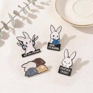 May include: Four enamel pins featuring white bunny designs. One bunny holds scissors, with the text "RUN WITH SCISSORS". Another reads a book with the text "SHHH... I'M READING". A third says "I'M NOT MEAN I'M HONEST". The fourth pin shows three bunnies.