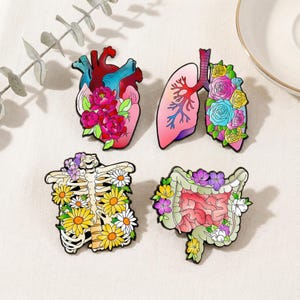 May include: Set of four enamel pins featuring anatomical designs: a heart, lungs, a rib cage, and intestines, each adorned with colorful floral arrangements. The pins are in shades of pink, red, blue, yellow, and white.