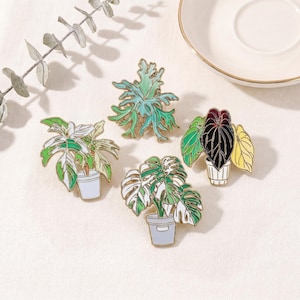 May include: Four enamel plant pins with gold trim. The pins feature detailed designs of various potted plants, including a Monstera, a fern, and other tropical foliage. Each plant is in a small, light-colored pot. The pins are set against a neutral background.