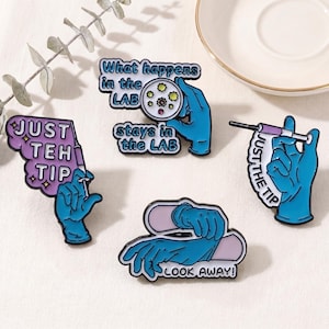 May include: Four enamel pins with medical themes. One pin reads "What happens in the LAB stays in the LAB" with a petri dish. Another says "JUST THE TIP" with a syringe. The other two pins feature gloved hands.