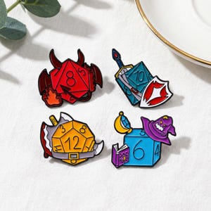 May include: Four enamel pins with a fantasy theme. The pins include a red die with horns and flames, a blue die with a sword and shield, a yellow die with a sword, and a blue cube with a purple wizard hat and book.