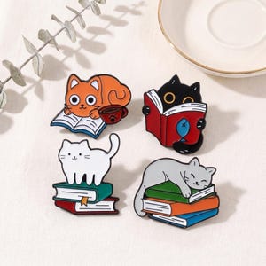 May include: Four cat-themed enamel pins. One orange cat reads a book with a coffee cup. A black cat reads a book with a fish. A white cat stands on books. A gray cat sleeps on books.