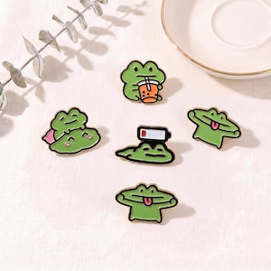 Könnte beinhalten: Ein Set aus fünf grünen Frosch-Emaille-Anstecknadeln. Ein Frosch hält einen Bubble Tea, einer hat ein Symbol für schwache Batterie auf dem Kopf, zwei strecken die Zunge heraus und ein Frosch sitzt auf einem anderen. Schwarze Konturen und bunte Details.