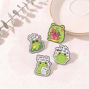 May include: Four enamel pins featuring green frogs with various expressions and accessories. One frog wears sunglasses with the text Just vibing, one holds a pink gift, one says It is What it is, and one holds a paper labelled PLAN with the text I had a plan.