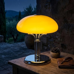May include: A yellow glass table lamp with a chrome base and stem. The lamp has a rounded, mushroom-shaped shade and sits on a round base. The lamp is on a wooden table with a small succulent plant and a leather-bound notebook.