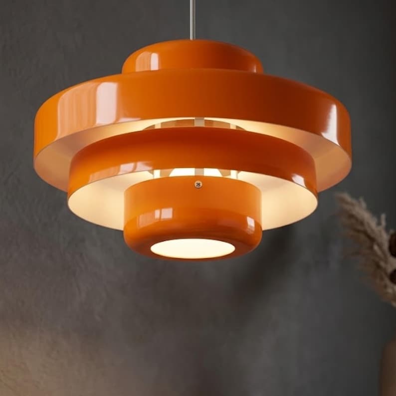 Pode incluir: Uma lumin&aacute;ria pendente laranja brilhante com um design em camadas composto por tr&ecirc;s c&uacute;pulas met&aacute;licas circulares conc&ecirc;ntricas. O interior de cada c&uacute;pula tem acabamento branco, refletindo a luz para baixo a partir do centro.