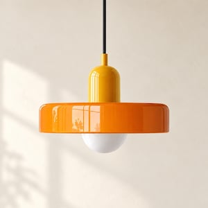 Op de afbeelding: Een hanglamp met een glanzende oranje schijfvormige kap, een felgeel cilindrisch bovendeel en een witte bolvormige lamp in het midden. De armatuur hangt aan een dun zwart snoer tegen een neutrale achtergrond.