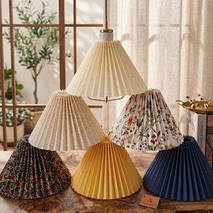 May include: A selection of pleated lampshades in diverse colours and patterns. Shades include cream, beige, yellow, and navy blue, with floral and patterned designs. The lampshades are displayed on a wooden surface, showcasing their different styles.