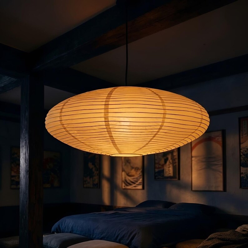 May include: An oval-shaped pendant light fixture with a warm, yellow glow. The lamp is made of a paper-like material with horizontal lines, suspended from a black cord. The light hangs above a bed in a room with dark blue walls and artwork.