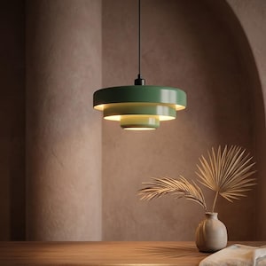 Pode incluir: Uma lumin&aacute;ria pendente de tr&ecirc;s n&iacute;veis na cor verde s&aacute;lvia, com tr&ecirc;s c&uacute;pulas circulares de metal conc&ecirc;ntricas de tamanhos decrescentes, suspensa por um fio preto fino, emitindo um brilho quente a partir do interior das camadas.