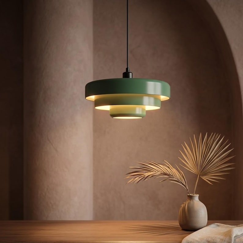 Pode incluir: Uma lumin&aacute;ria pendente de tr&ecirc;s n&iacute;veis na cor verde s&aacute;lvia, com tr&ecirc;s c&uacute;pulas circulares de metal conc&ecirc;ntricas de tamanhos decrescentes, suspensa por um fio preto fino, emitindo um brilho quente a partir do interior das camadas.