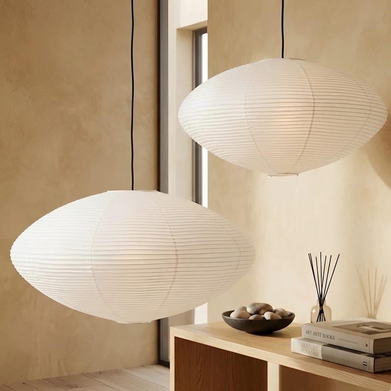 May include: Two white, oval-shaped pendant lights hang from black cords. The lamps have a ribbed, paper-like texture. The lights are suspended in a room with a neutral color palette, suggesting a modern or minimalist design aesthetic.