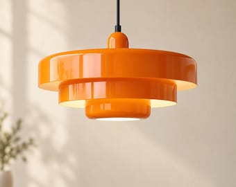 Pode incluir: Uma luminária pendente laranja brilhante com um design circular de três níveis e acabamento lustroso. A peça é suspensa por um fio preto fino e emite uma luz branca suave a partir da parte inferior de cada anel metálico concêntrico.