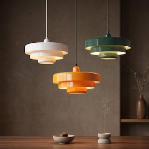 Pode incluir: Tr&ecirc;s candeeiros suspensos de tr&ecirc;s n&iacute;veis em branco, laranja e verde escuro, cada um com um acabamento liso e brilhante e um design circular de v&aacute;rias camadas. Estes candeeiros est&atilde;o suspensos por cabos finos e emitem um brilho quente.