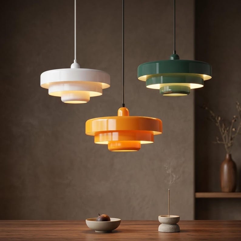 Pode incluir: Tr&ecirc;s candeeiros suspensos de tr&ecirc;s n&iacute;veis em branco, laranja e verde escuro, cada um com um acabamento liso e brilhante e um design circular de v&aacute;rias camadas. Estes candeeiros est&atilde;o suspensos por cabos finos e emitem um brilho quente.