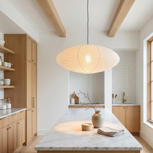 May include: A modern kitchen with light wood cabinetry and a marble countertop island. A large, oval-shaped paper lantern hangs from the ceiling, providing soft lighting. The room features a neutral color palette with natural elements.