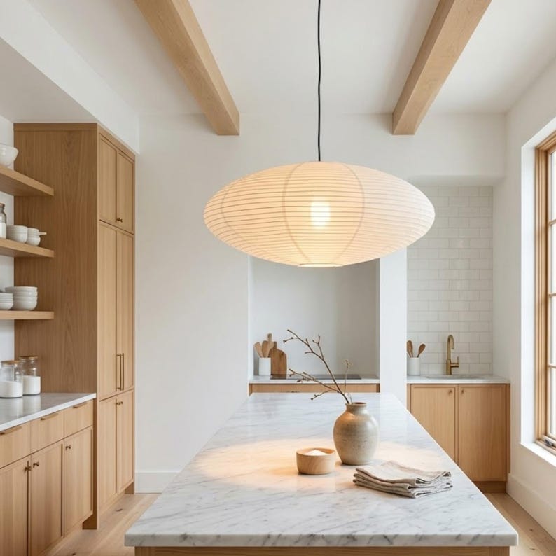 May include: A modern kitchen with light wood cabinetry and a marble countertop island. A large, oval-shaped paper lantern hangs from the ceiling, providing soft lighting. The room features a neutral color palette with natural elements.