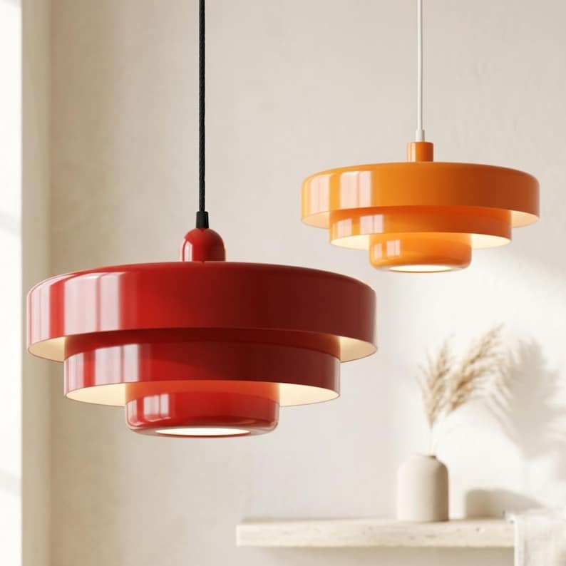 Pode incluir: Duas lumin&aacute;rias pendentes em camadas com acabamento brilhante. A luz em primeiro plano &eacute; vermelho-escura com um fio preto, enquanto a luz ao fundo &eacute; laranja brilhante com um fio branco. Ambas apresentam um design circular empilhado.