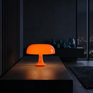 Pode incluir: Um candeeiro de mesa cor de laranja brilhante em forma de cogumelo com um acabamento liso e brilhante. O candeeiro apresenta um abajur largo e arredondado em forma de cúpula, suportado por uma base central, emitindo um brilho quente.