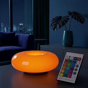 May include: A vibrant orange, ring-shaped table lamp with a black base. A remote control with colour options is positioned beside the lamp. The lamp is illuminated, emitting a warm light. The background features a dark blue wall and a plant in a vase.