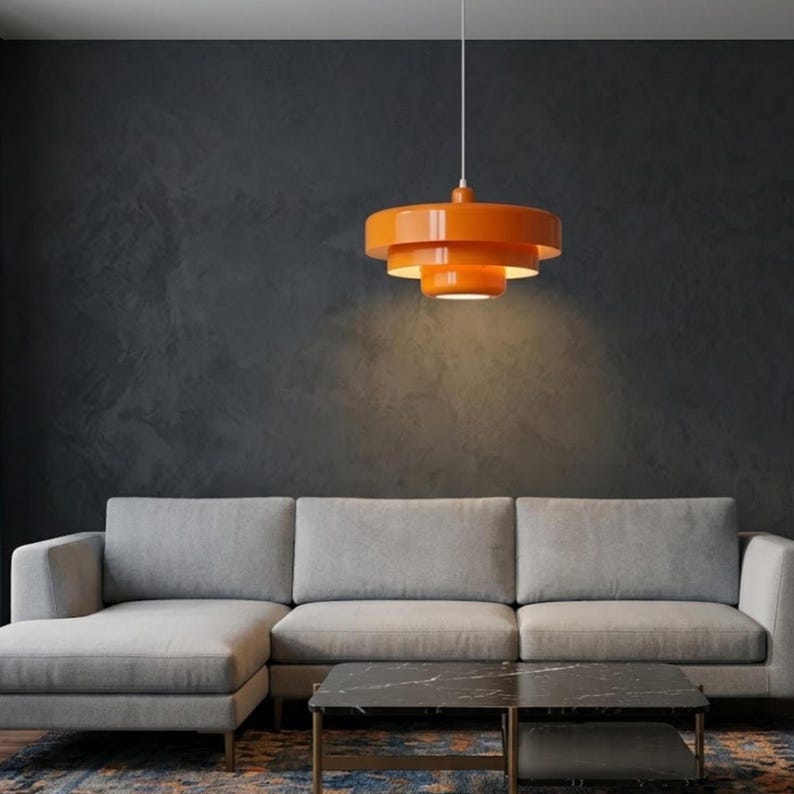 Pode incluir: Uma lumin&aacute;ria pendente de cor laranja brilhante com um design circular em camadas, apresentando tr&ecirc;s an&eacute;is empilhados de tamanhos variados. A l&acirc;mpada est&aacute; suspensa por um fio branco fino e emite uma luz quente direcionada para baixo.