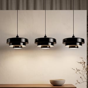 Pode incluir: Tr&ecirc;s candeeiros suspensos pretos com um design circular de v&aacute;rios n&iacute;veis, pendurados por cabos pretos finos. Cada candeeiro apresenta um acabamento brilhante no exterior e um interior branco, projetando luz para baixo.