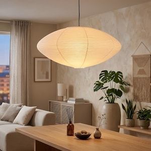 May include: A large, oval-shaped pendant light fixture made of a cream-colored, ribbed paper material hangs from the ceiling. The light illuminates a living room with a neutral color palette, including a wooden table, sofa, and decorative plants.