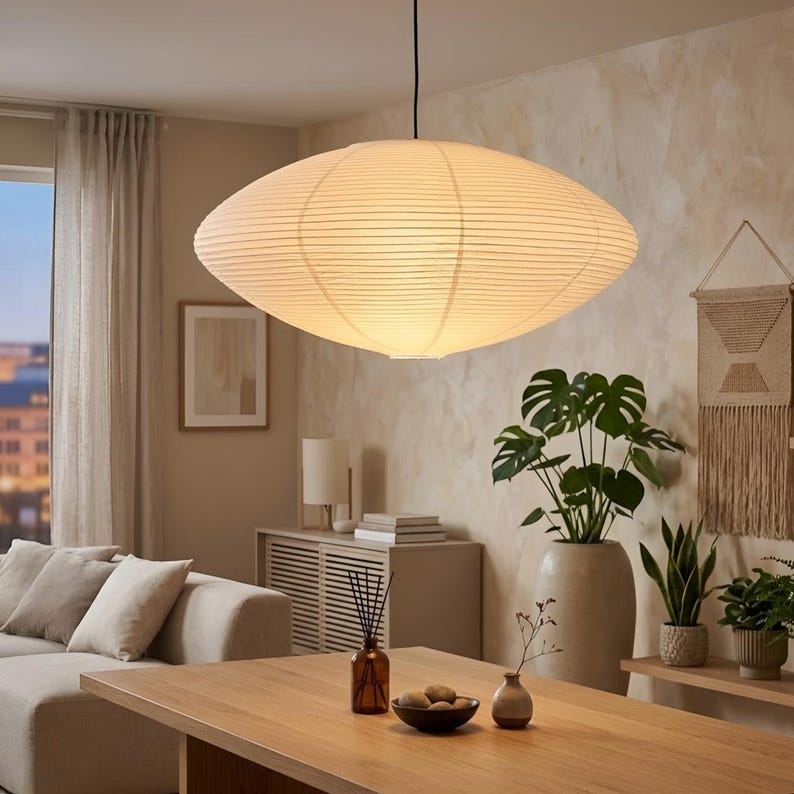 May include: A large, oval-shaped pendant light fixture made of a cream-colored, ribbed paper material hangs from the ceiling. The light illuminates a living room with a neutral color palette, including a wooden table, sofa, and decorative plants.