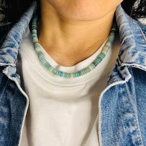 May include: A beaded necklace with a mix of light blue, green, and white round beads. The necklace is worn over a white t-shirt and a blue denim jacket. The beads appear to be small and evenly sized, creating a delicate look.