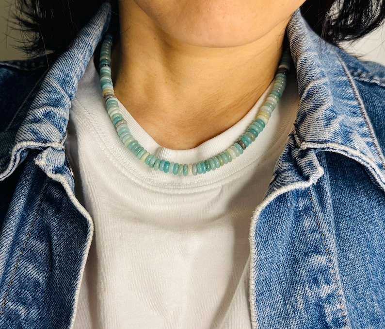 May include: A beaded necklace with a mix of light blue, green, and white round beads. The necklace is worn over a white t-shirt and a blue denim jacket. The beads appear to be small and evenly sized, creating a delicate look.