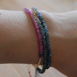 May include: Three layered bracelets with pink, blue, and gray gemstone beads. The bracelets are on a person's wrist.