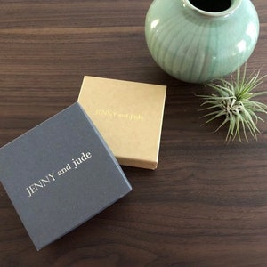May include: Two small square boxes with the text "JENNY and jude" printed in gold on the top. The box on the left is gray and the box on the right is brown.