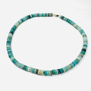May include: A turquoise beaded necklace with a gold clasp. The necklace is made of small, round beads in shades of turquoise, green, and gray. The beads are strung together to form a circular shape. The necklace is displayed on a white background.
