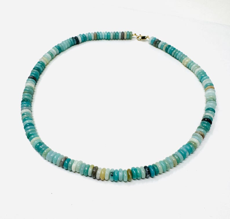 May include: A turquoise beaded necklace with a gold clasp. The necklace is made of small, round beads in shades of turquoise, green, and gray. The beads are strung together to form a circular shape. The necklace is displayed on a white background.