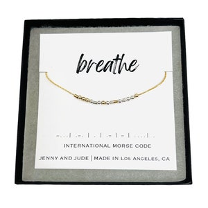 May include: A gold-toned necklace with a Morse code design, presented in a black box. The necklace is strung with small beads and bars. The card behind the necklace reads "breathe" and "International Morse Code". The text "JENNY AND JUDE | MADE IN LOS ANGELES, CA" is also visible.