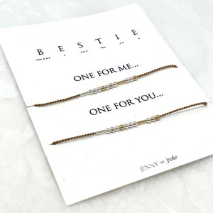 May include: Two brown friendship bracelets with silver and gold beads and a small metal bar. The bracelets are on a white card with the text "BESTIE... ONE FOR ME... ONE FOR YOU... JENNY jude".