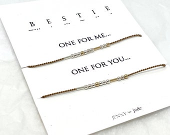 Puede incluir: Dos pulseras de amistad marrones con cuentas plateadas y doradas y una pequeña barra de metal. Las pulseras están sobre una tarjeta blanca con el texto "BESTIE... ONE FOR ME... ONE FOR YOU... JENNY jude".