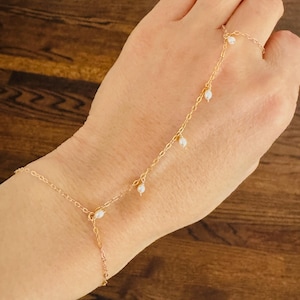 Könnte beinhalten: Ein goldenes Kettenarmband mit kleinen weißen Perlen, die daran hängen. Das Armband ist so konzipiert, dass es am Handgelenk und am Finger getragen werden kann.