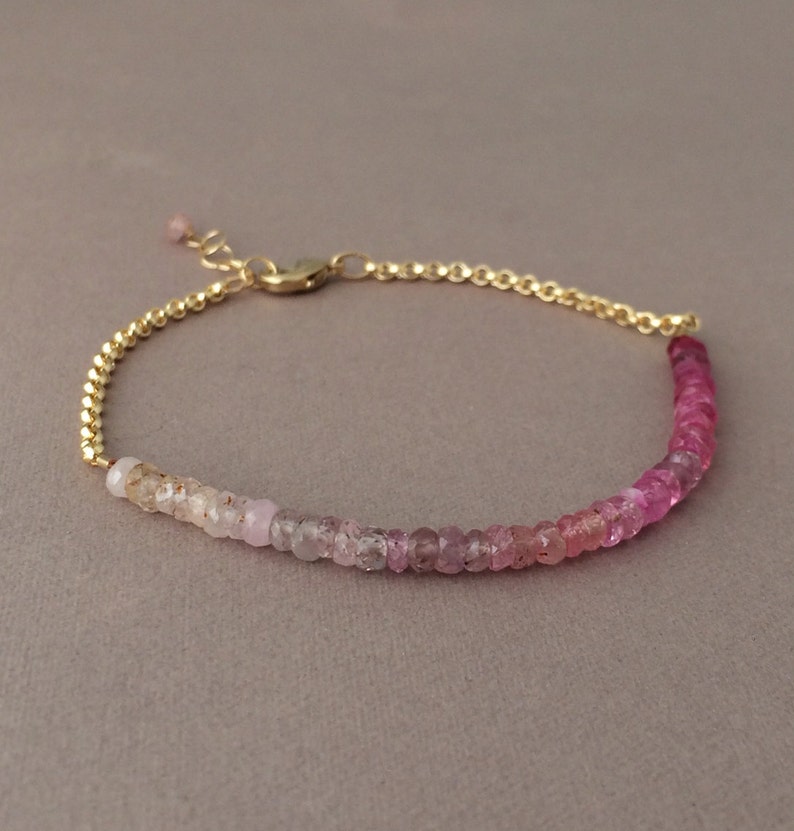 May include: A delicate gold chain bracelet with a gradient of pink and white gemstones. The bracelet is made with small, round beads that are strung together in a continuous line.