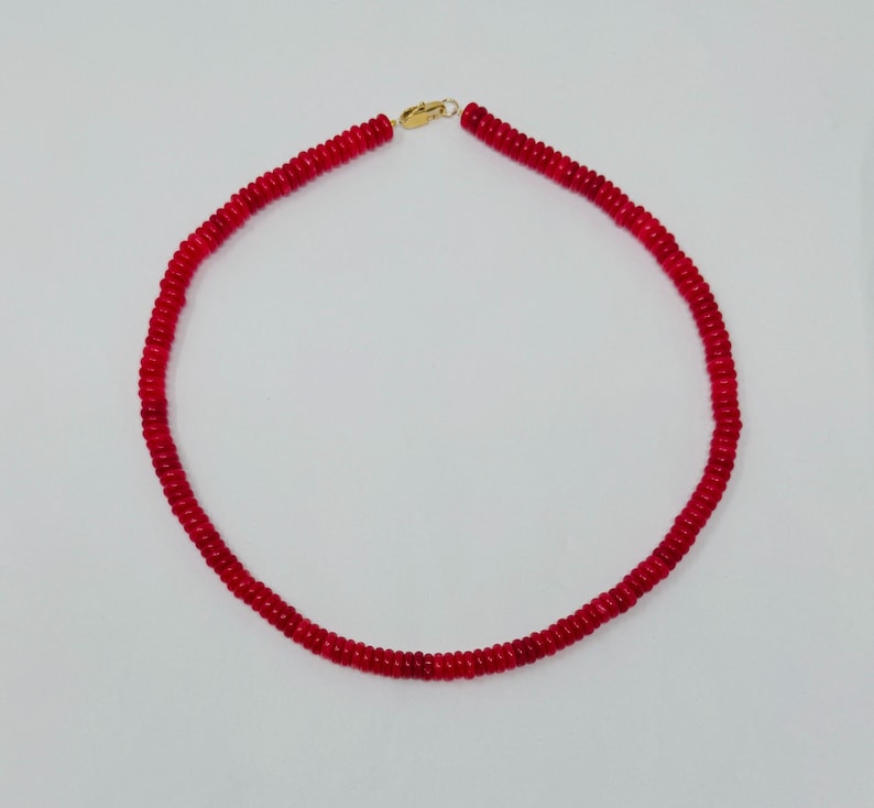 May include: A red beaded necklace with a gold-colored clasp. The necklace is composed of small, round, red beads strung together to form a circular shape. The clasp is a simple hook and eye closure.