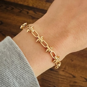 May include: A gold chain bracelet with pointed spikes. The bracelet is on a person's wrist.