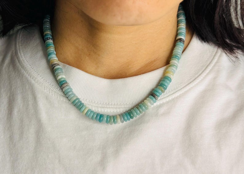 May include: A beaded necklace with a mix of light blue, white, and gray beads. The necklace is worn around the neck and rests on a white t-shirt. The beads are small and round, creating a delicate and colorful accessory.