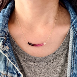 May include: A delicate gold necklace featuring a gradient of colorful beads, transitioning from dark to light shades of pink and white. The necklace is worn against a gray top and a denim jacket.