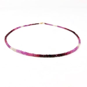 May include: A delicate necklace composed of small, faceted gemstones in shades of pink, purple, and white. The necklace has a gold clasp and is displayed against a white background. The gemstones transition from dark to light colors.