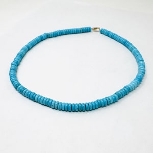 May include: A turquoise beaded necklace with a gold-colored clasp. The necklace is composed of small, round, light blue beads. The beads are strung together to form a circular shape. The necklace is displayed on a white background.