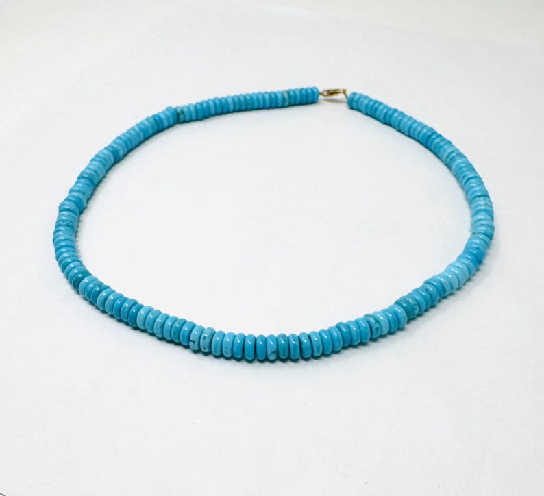 May include: A turquoise beaded necklace with a gold-colored clasp. The necklace is composed of small, round, light blue beads. The beads are strung together to form a circular shape. The necklace is displayed on a white background.