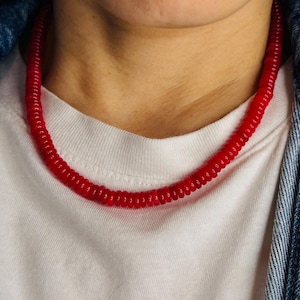 May include: A vibrant red necklace composed of small, connected cylindrical beads. The necklace is worn against a white t-shirt and a denim jacket. The necklace is a bold accessory.