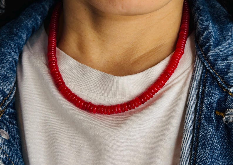 May include: A vibrant red necklace composed of small, connected cylindrical beads. The necklace is worn against a white t-shirt and a denim jacket. The necklace is a bold accessory.